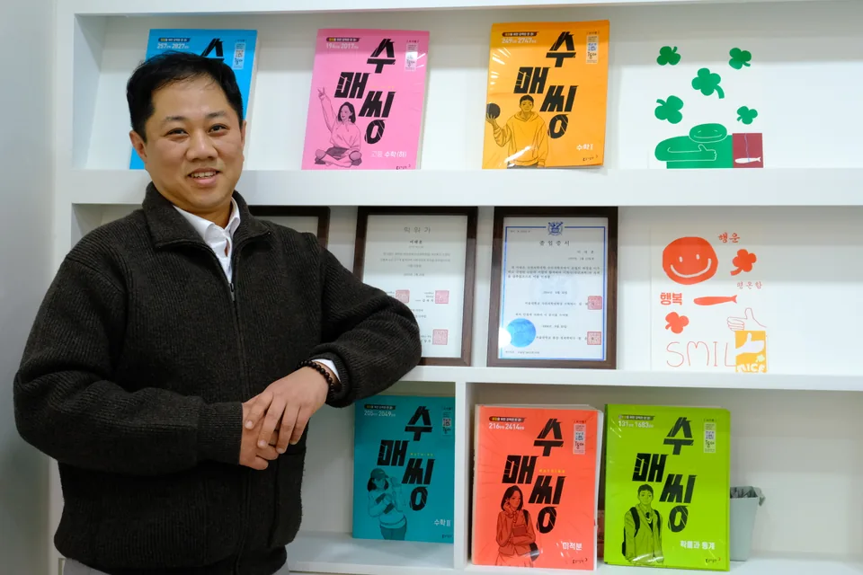 이태훈 원장이 「수매씽」 교재와 표창장이 진열된 서가 앞에 서 있는 모습